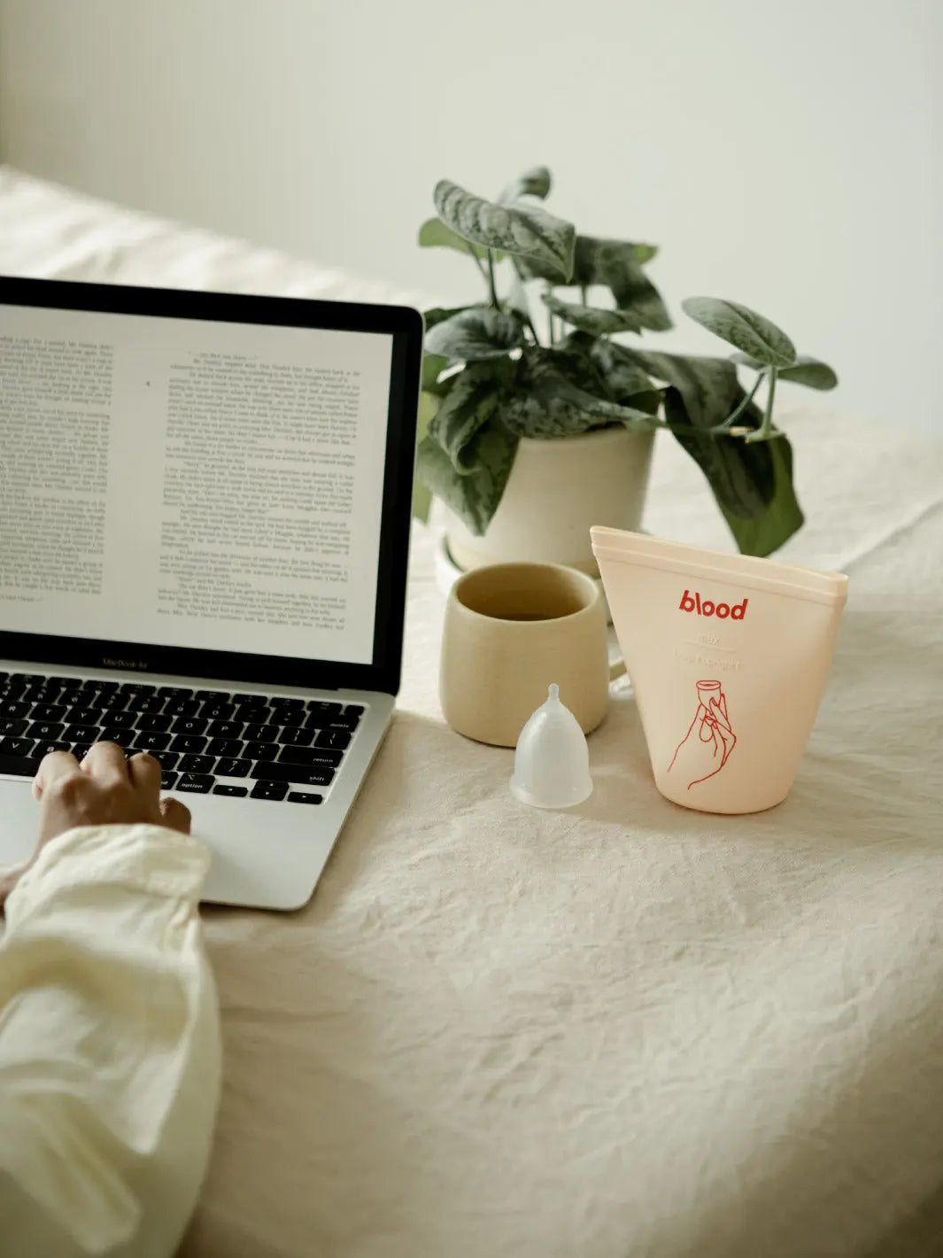 a girl is sitting at a table using her laptop. there is a menstrual cup and sanitising pouch on the table.
